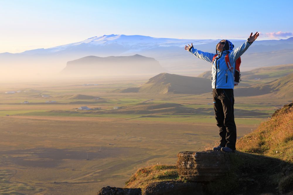 Hiker with outstretched arms celebrating at sunrise, with views over southern Iceland’s farmlands and distant snow-covered peaks