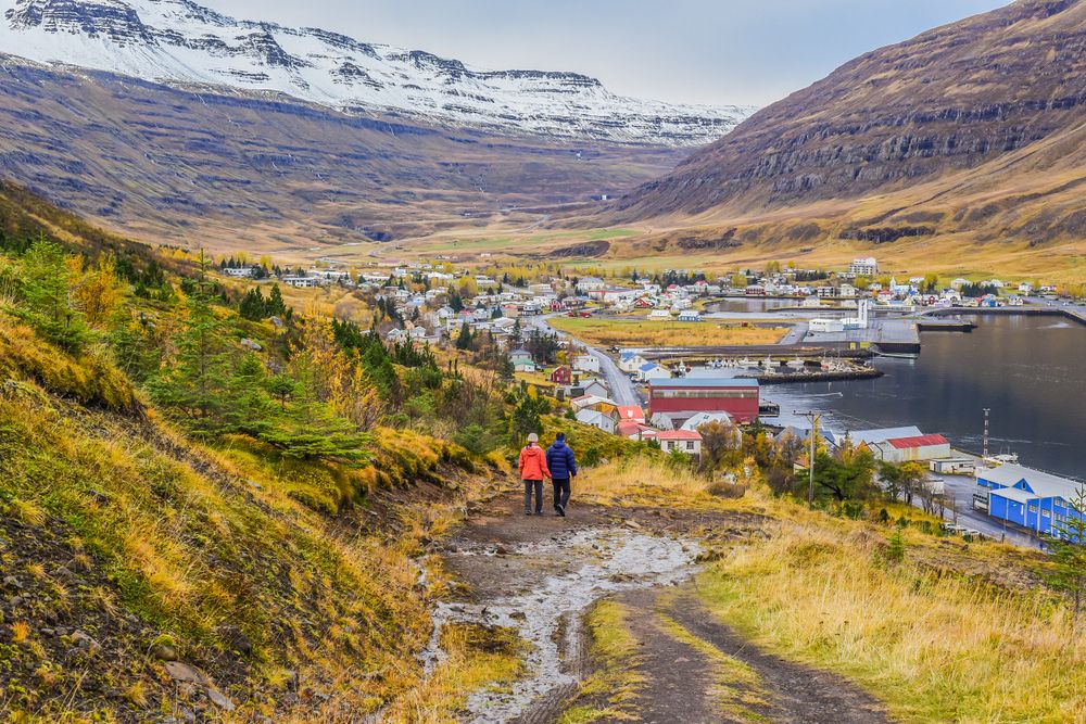 Two people walking on a trail overlooking Seyðisfjörður village with autumn colors and snow-dusted mountains in the background