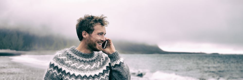  Smiling man in traditional Icelandic sweater making a phone call by the coast on a cloudy day.