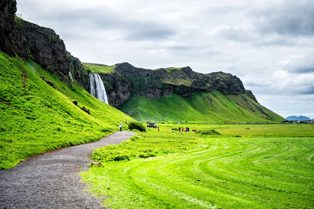 cenic walking path toward Seljalandsfoss waterfall with tourists and lush green cliffs.