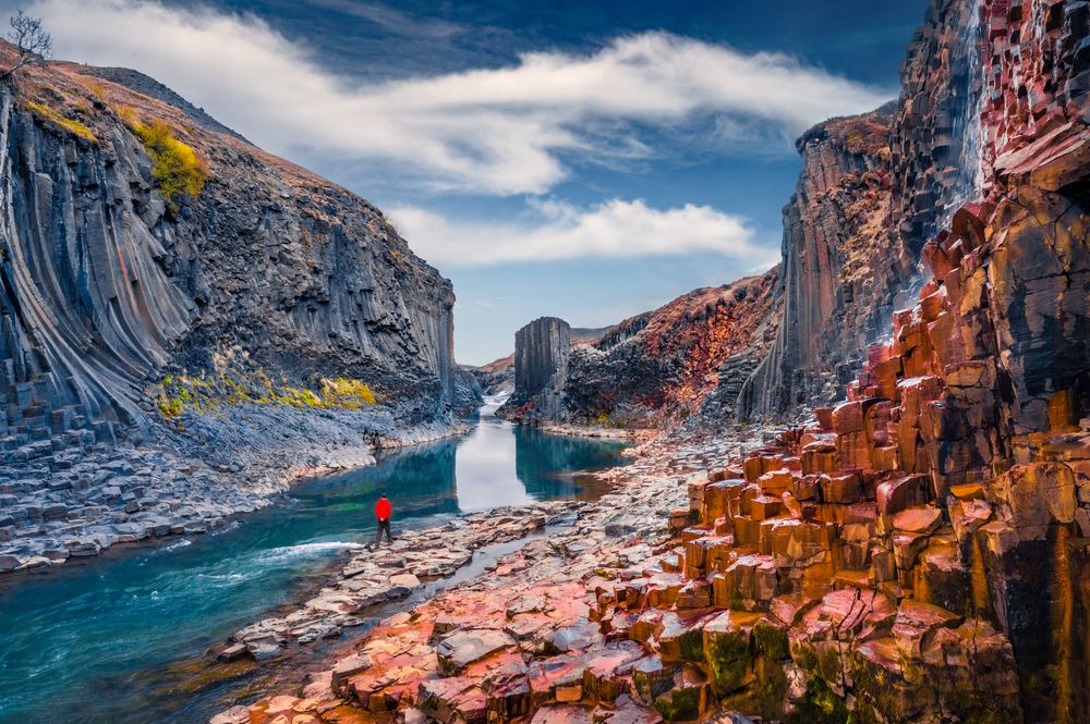 Person standing beside the blue river at Studlagil Canyon surrounded by tall basalt columns and vivid autumn colors