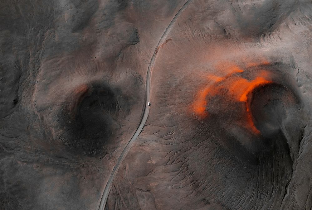 Aerial view of a single car driving on a winding road through Iceland’s surreal volcanic landscape, with dark earth tones and vibrant orange streaks