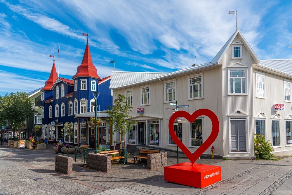 Red heart sculpture on a pedestrian street in downtown Akureyri, surrounded by colorful historic buildings and summer flowers
