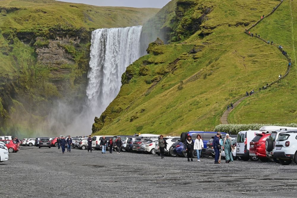 Crowded parking lot at Skógafoss waterfall with many parked cars and tourists.