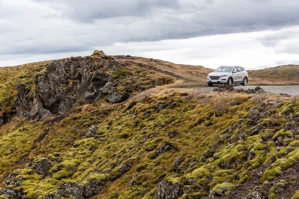 A silver SUV parked on a narrow road surrounded by mossy cliffs and dramatic Icelandic terrain under overcast skies