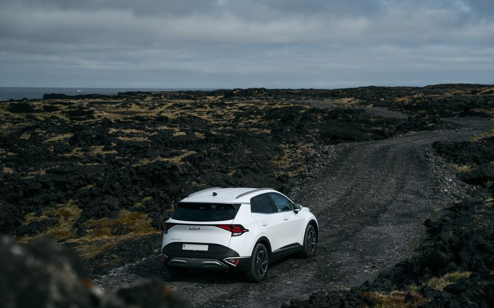 White vehicle driving through black lava fields along a gravel road in Iceland, with the ocean barely visible in the distance.
