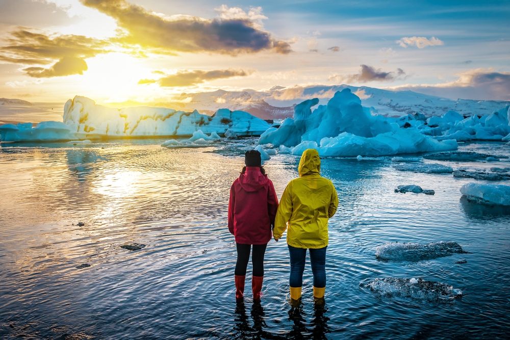 Two people in red and yellow jackets standing hand-in-hand at Jökulsárlón Glacier Lagoon in Iceland during sunset.