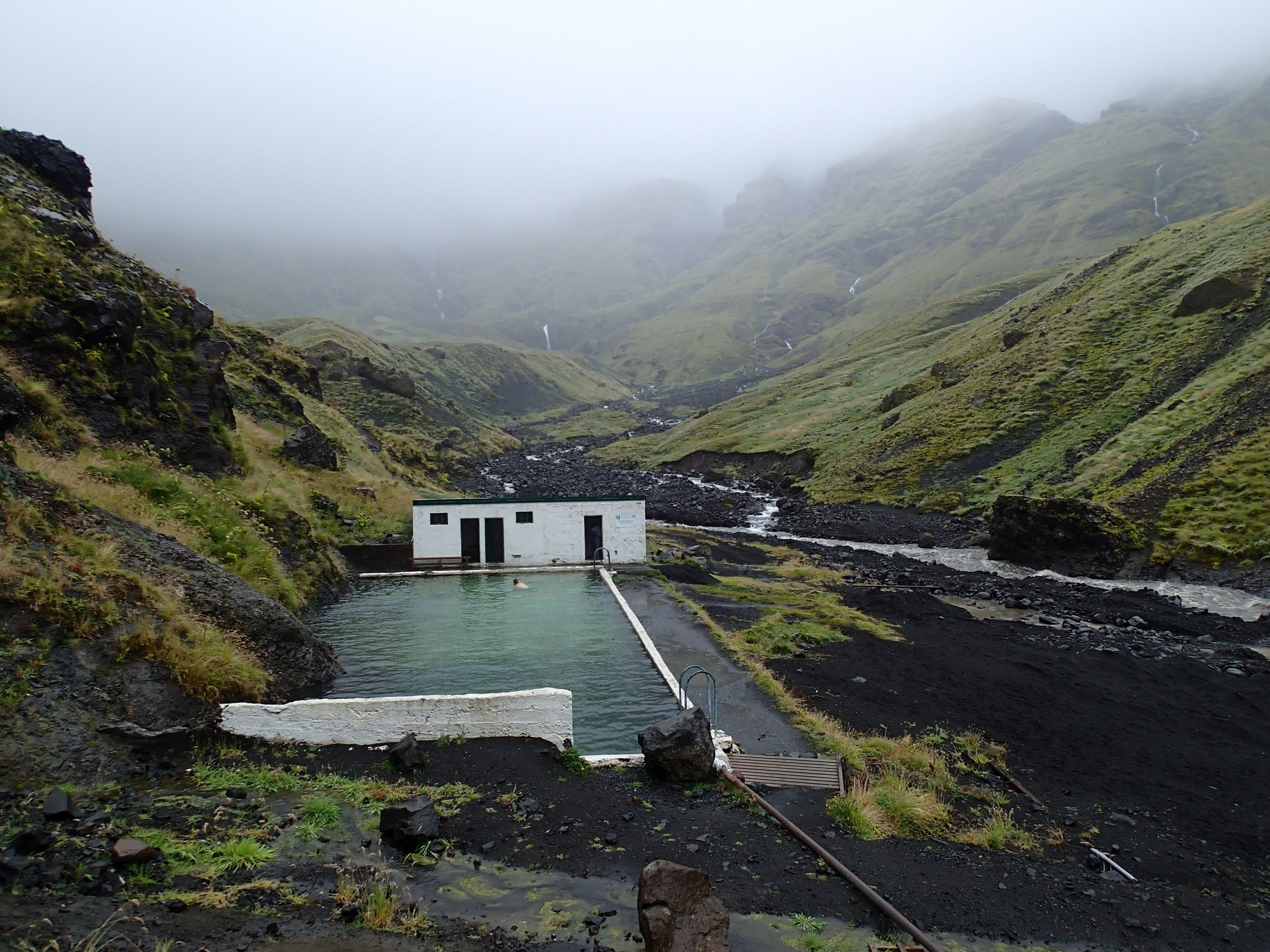The historic Seljavallalaug pool nestled in a misty green valley, surrounded by volcanic mountains and rivers